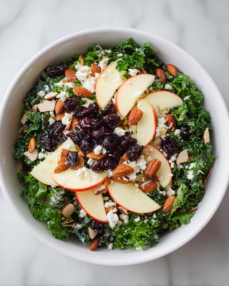 A white bowl filled with a fresh kale salad showing about three layers: the bottom layer is dark green kale leaves with a curly texture, the middle layer has thin, pale red and white apple slices placed evenly around the salad, and the top layer consists of dark dried cherries, light brown toasted almond slices, and small white crumbles of cheese scattered across. The bowl sits on a white marbled surface. photo taken with an iphone --ar 4:5 --v 7