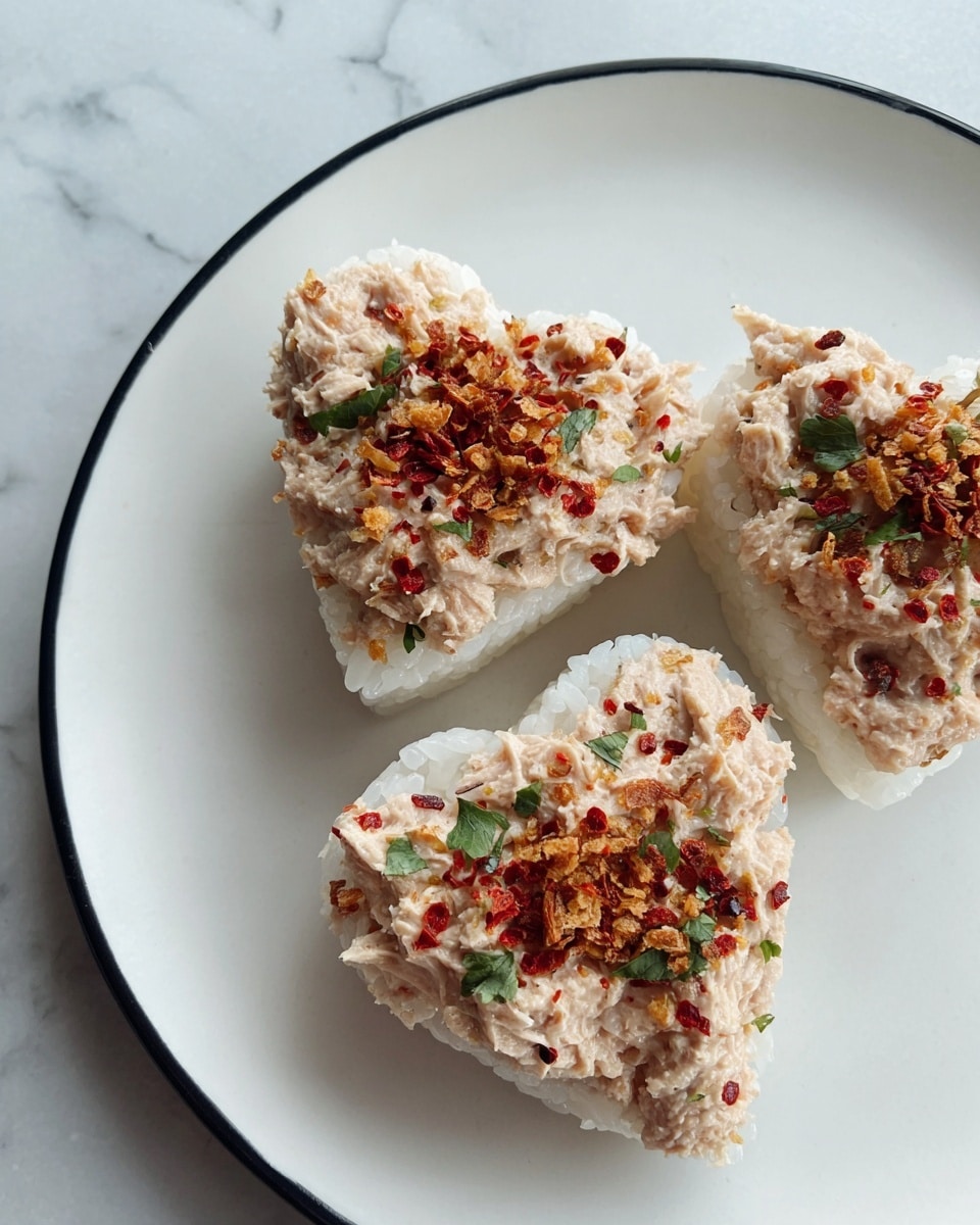 The image shows three heart-shaped rice cakes on a white plate, each topped with a creamy, light pink tuna mixture. The rice base is white with a sticky texture, slightly translucent around the edges. The tuna topping covers each rice cake thickly, sprinkled with red chili flakes, crispy fried bits, and small green herb leaves, adding color and texture contrasts. The plate sits on a white marbled surface. photo taken with an iphone --ar 4:5 --v 7