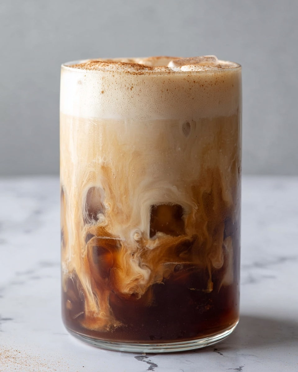A clear glass filled with a three-layered iced coffee drink stands on a white marbled surface. The bottom layer is a dark brown coffee base with visible ice cubes. Above it, a middle layer features swirling light brown and creamy beige colors, showing the mixing of coffee and cream. The top layer is a thick, foamy, light beige cream that covers the surface with a smooth texture and a dusting of light brown powder on top. photo taken with an iphone --ar 4:5 --v 7